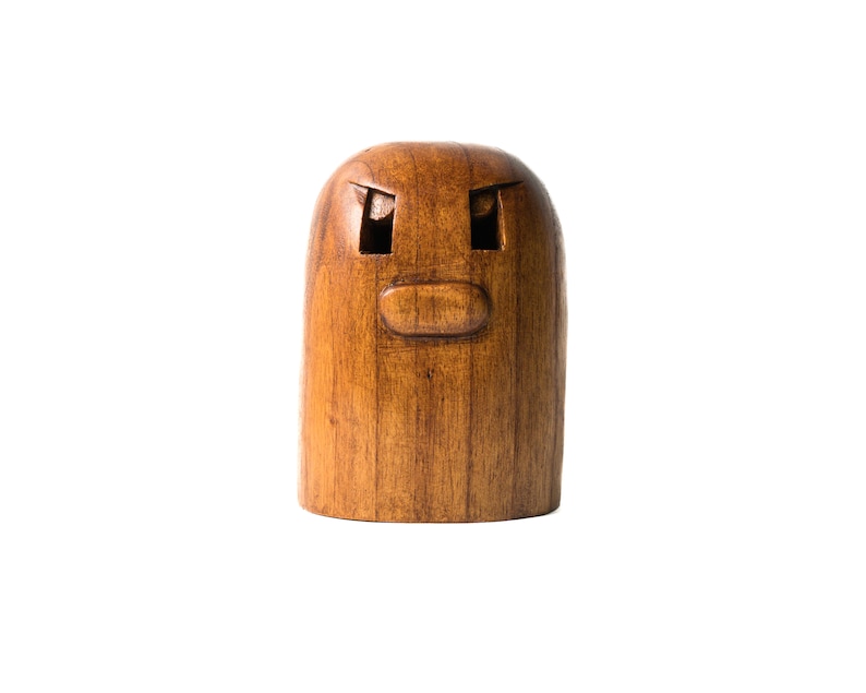 May include: A wooden figurine with a carved face, featuring two eyes and a mouth. The figurine is brown and has a smooth, polished finish.