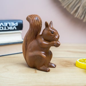 May include: A wooden squirrel figurine with a bushy tail, sitting on a light brown wooden surface. The squirrel is holding a nut in its paws.
