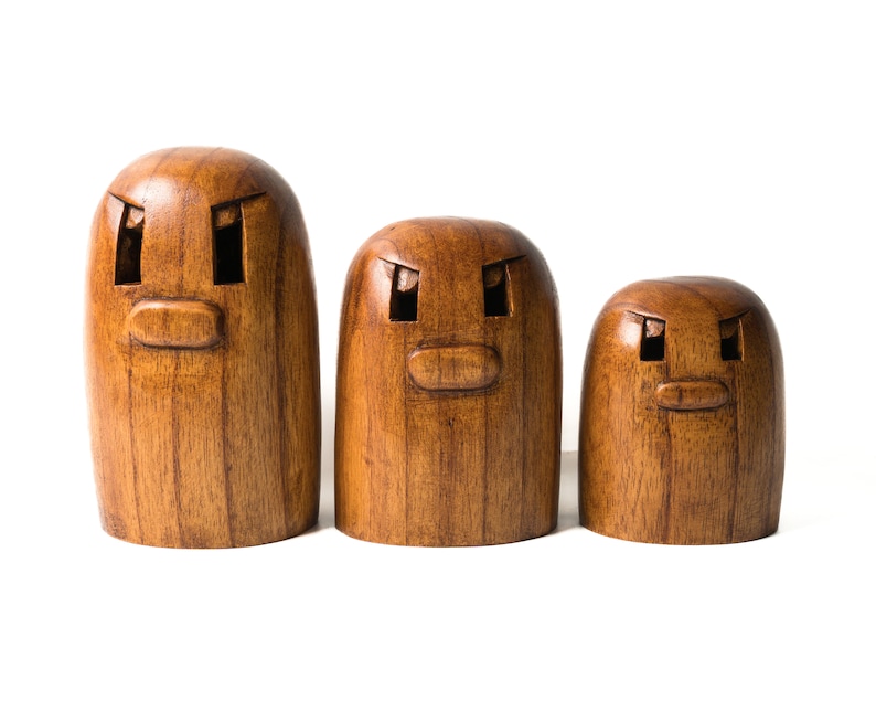 May include: Three wooden figurines with carved faces. The figurines are brown and have a simple design. The largest figurine is on the left, the middle figurine is in the center, and the smallest figurine is on the right.