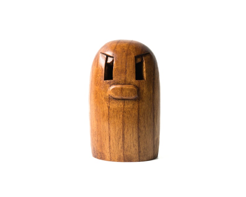 May include: A wooden figurine with a carved face, featuring two eyes and a mouth. The figurine is brown and has a smooth, polished finish.