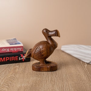 May include: A wooden carving of a dodo bird, a flightless bird native to Mauritius. The carving is brown and has a natural wood finish. The dodo is standing on a wooden base.