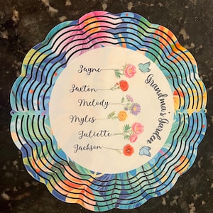 May include: A round, layered garden decoration with a floral and leaf pattern in shades of blue, pink, and orange. The center features names and the text "Grandma's Garden" with flower illustrations and butterfly accents.