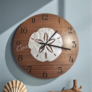 May include: A round wooden wall clock with a white sand dollar design in the center. The clock has black hands and black numerals. Decorative seashells and driftwood are displayed below the clock.