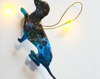 May include: A dachshund-shaped ornament featuring a glossy finish with a swirling galaxy pattern in shades of black, deep blue, and teal, accented with silver glitter. A thin gold cord is attached to the top for hanging. The number 2 is visible.