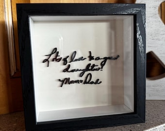 Op de afbeelding: Een zwart omrandde schaduwdoos met de tekst "Lots of love to our daughter! Mom & Dad" in cursief. De achtergrond is wit.