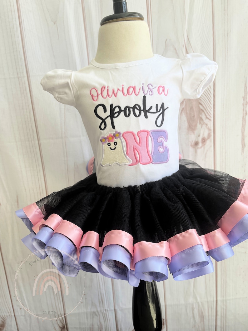 May include: A white onesie with the text "Olivia is a Spooky One" in pink and black lettering. A ghost with a flower crown is embroidered on the onesie. The onesie is paired with a black tulle tutu with pink and purple ribbon trim.