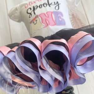 May include: A white onesie with the text "Olivia is a Spooky One" in pink and purple lettering. A ghost with a flower crown is embroidered on the onesie. The onesie is paired with a black tulle skirt with pink and purple satin ribbon trim.