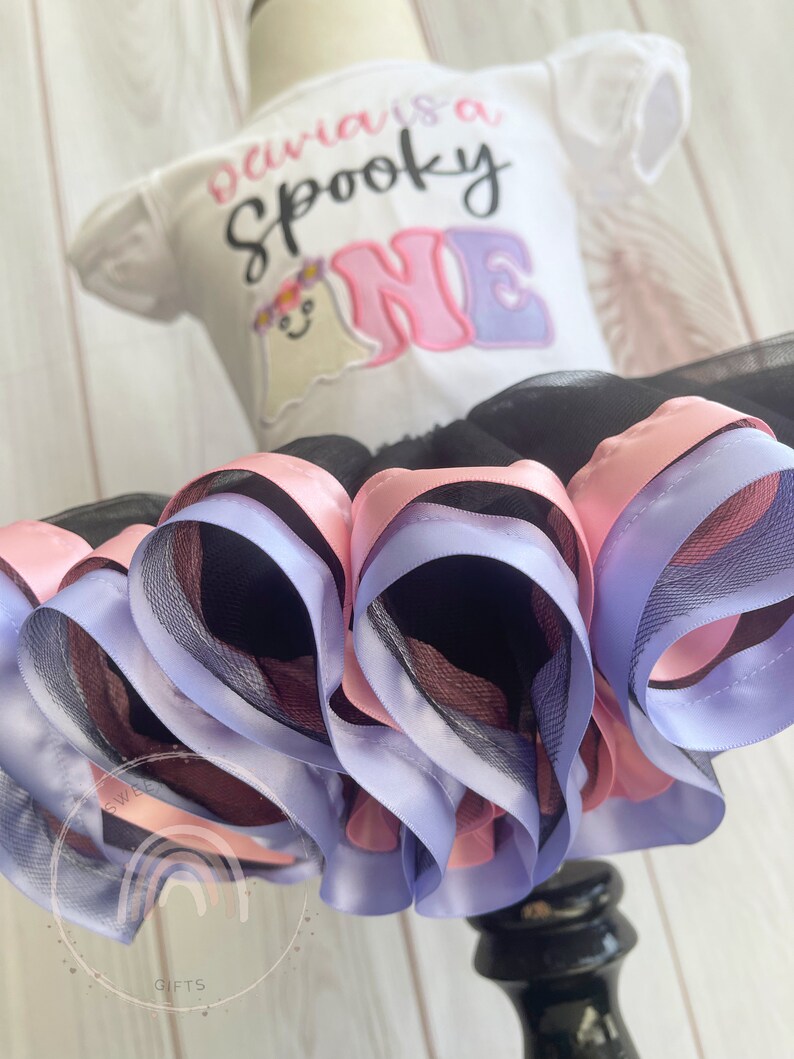 May include: A white onesie with the text "Olivia is a Spooky One" in pink and purple lettering. A ghost with a flower crown is embroidered on the onesie. The onesie is paired with a black tulle skirt with pink and purple satin ribbon trim.