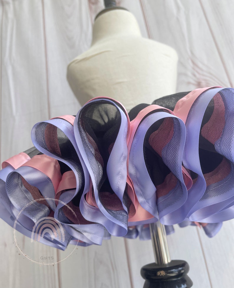 May include: A black tulle tutu skirt with layers of pink and lavender satin ribbon. The skirt is on a white dress form.