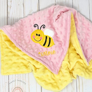 May include: A pink and yellow minky baby blanket with a bee and the name Cortnie embroidered on it. The blanket is made by Sweet Threads Gifts.