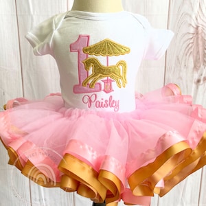 May include: A pink tutu with gold ribbon trim and a white onesie with a pink number one and a gold carousel horse design. The onesie is personalized with the name "Paisley".