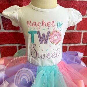 May include: A white short-sleeved shirt with the text "Rachee is TWO Sweet" embroidered on the front. The number two is decorated with sprinkles and a pink and white donut. The shirt is paired with a multi-colored tulle skirt.
