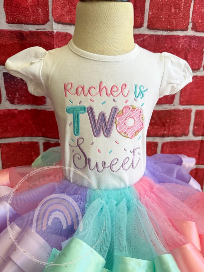 May include: A white short-sleeved shirt with the text "Rachee is TWO Sweet" embroidered on the front. The number two is decorated with sprinkles and a pink and white donut. The shirt is paired with a multi-colored tulle skirt.
