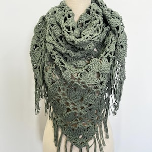 May include: A sage green crochet shawl with a triangular shape and fringe detailing. The shawl features an open, lacy design and is displayed on a mannequin. The fringe hangs from the bottom edge.