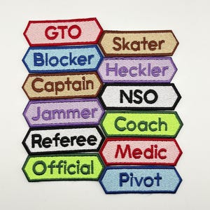 Peut inclure: Un ensemble de 10 patchs thermocollants colorés avec différents mots, dont "GTO", "Skater", "Blocker", "Heckler", "Captain", "NSO", "Jammer", "Coach", "Referee", "Medic", "Official", et "Pivot".
