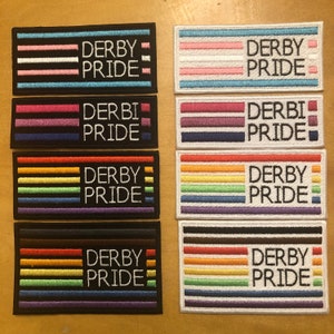 Op de afbeelding: Acht geborduurde patches met de tekst "Derby Pride" in witte letters op een zwarte achtergrond. Elke patch heeft een ander kleurenschema, waaronder regenboog-, transgender- en lesbische pride-vlaggen.