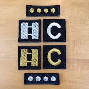 Könnte beinhalten: Vier schwarze Stoffpatches mit weißen und goldenen Stickereien. Zwei Patches zeigen den Buchstaben "C" und zwei Patches zeigen den Buchstaben "H" mit zwei horizontalen Balken. Ein kleiner schwarzer Stoffpatch mit drei goldenen Punkten und ein kleiner schwarzer Stoffpatch mit drei weißen Punkten.
