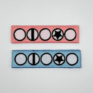 Peut inclure: Deux rangées de patchs brodés avec cinq cercles chacun. La rangée du haut est rose avec des cercles blancs et une étoile noire au centre du quatrième cercle. La rangée du bas est bleu clair avec des cercles blancs et une étoile noire au centre du quatrième cercle.