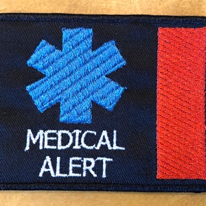 Może przedstawiać: Czarna naszywka haftowana z niebieską gwiazdą życia, napisem "MEDICAL ALERT" w kolorze białym i czerwonym paskiem po prawej stronie.
