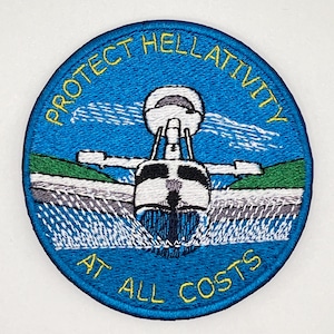 May include: A circular embroidered patch with a blue background featuring a white seaplane taking off from water. The text "PROTECT HELLATIVITY" is arched at the top and "AT ALL COSTS" at the bottom in yellow thread. Green hills are visible.