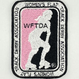 May include: A white, black and pink embroidered patch featuring a silhouette of a roller derby skater. The patch has the text "Women's Flat Track Derby Association" and "WFTDA" in a white font.