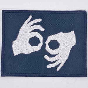 Può includere: Una toppa blu scuro con mani ricamate in bianco che formano il simbolo della lingua dei segni italiana per "va bene".