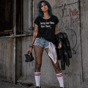 May include: A black t-shirt with the white text Keep your Chaos Over There, blue denim shorts, black fishnet stockings, white knee-high socks with red stripes, a white patterned bandana, and a black leather jacket held in one hand.
