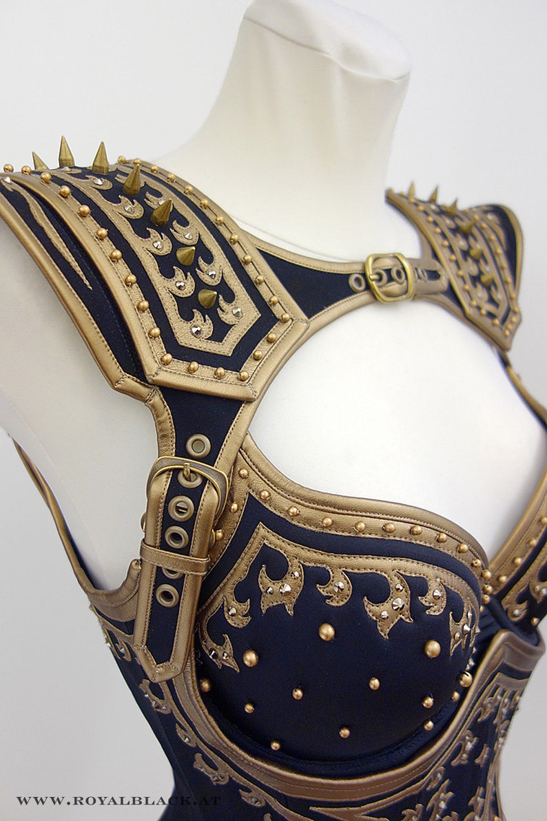 May include: A black and gold leather corset with a decorative design and spikes. The corset has a strap that goes over the shoulder and a buckle in the front.