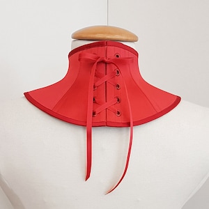 May include: Red satin choker with a lace-up back and ribbon ties. The choker is on a white mannequin.