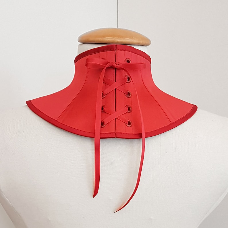 May include: Red satin choker with a lace-up back and ribbon ties. The choker is on a white mannequin.