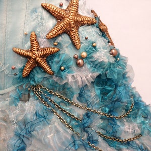 Puede incluir: Un cors&eacute; de tela azul y blanco con adornos de estrellas de mar doradas y un detalle de cadena dorada. La tela est&aacute; en capas y fruncida, creando un look caprichoso y elegante.