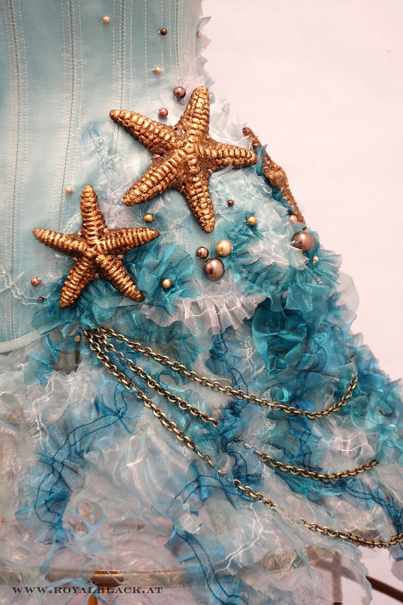 Puede incluir: Un cors&eacute; de tela azul y blanco con adornos de estrellas de mar doradas y un detalle de cadena dorada. La tela est&aacute; en capas y fruncida, creando un look caprichoso y elegante.