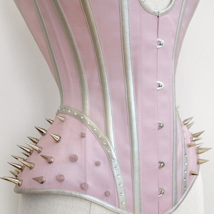 May include: A pink and silver corset with a pointed, studded design. The corset has a busk closure and is made of a satin-like fabric.