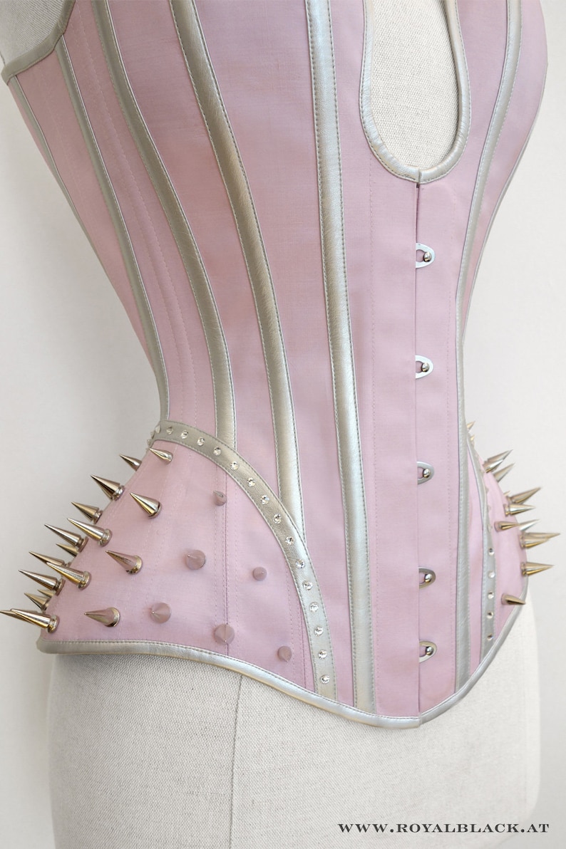 May include: A pink and silver corset with a pointed, studded design. The corset has a busk closure and is made of a satin-like fabric.