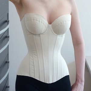May include: A white strapless corset with boning and a pointed bottom. The corset is made of a smooth fabric and has a classic design.