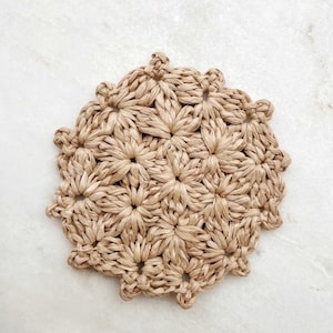 May include: A circular, beige woven coaster with a floral pattern. The coaster is crafted from natural fibres and displays a repeating design of flower-like forms. It is designed to protect surfaces from heat and moisture.