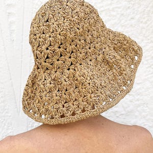 Könnte beinhalten: Ein sonnenhut aus gewebtem Stroh in hellem Beige. Der Hut hat ein Blumenmuster auf der Krone und dem Rand. Der Rand ist gewellt. Er dient als Sonnenschutz.