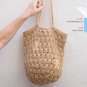 Può includere: Una borsa tote beige all'uncinetto con un motivo floreale. La borsa ha due manici ed è tenuta contro un muro bianco. Il testo sul lato destro dell'immagine recita "LEVEL INTERMEDIATE SIZE MULTIPLE."