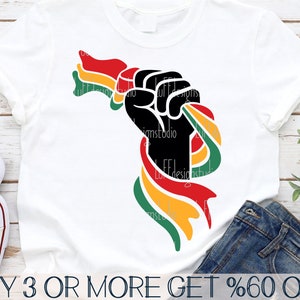 Peut inclure: Un poing noir levé en l'air, avec un ruban rouge, jaune et vert enroulé autour. Le ruban est un symbole du drapeau panafricain. L'image est sur un t-shirt blanc.