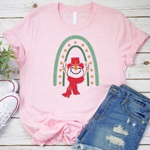 May include: A pink t-shirt with a green and gold rainbow design and a snowman in the center. The snowman is wearing a red hat and scarf.