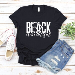 May include: Black t-shirt with a white graphic that says "Black is beautiful" with an outline of the continent of Africa in the word "Black".