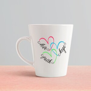 May include: White ceramic mug with a design of three overlapping hearts in pink, blue, and green. The words "love", "hope", and "faith" are written inside the hearts.