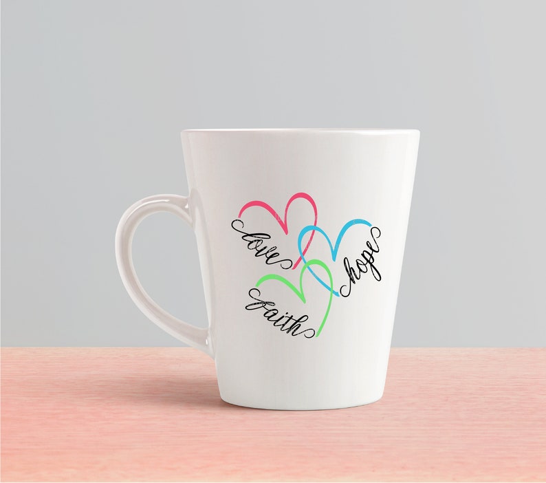 May include: White ceramic mug with a design of three overlapping hearts in pink, blue, and green. The words "love", "hope", and "faith" are written inside the hearts.