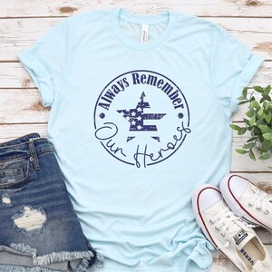 May include: Light blue t-shirt with a distressed navy blue circle graphic featuring a star with an American flag design and the text "Always Remember Our Heroes".