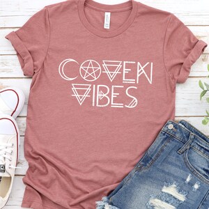 May include: A mauve t-shirt with the text "COVEN VIBES" in white. The text is stylized with geometric shapes and symbols, including a crescent moon, a pentagram, and triangles.