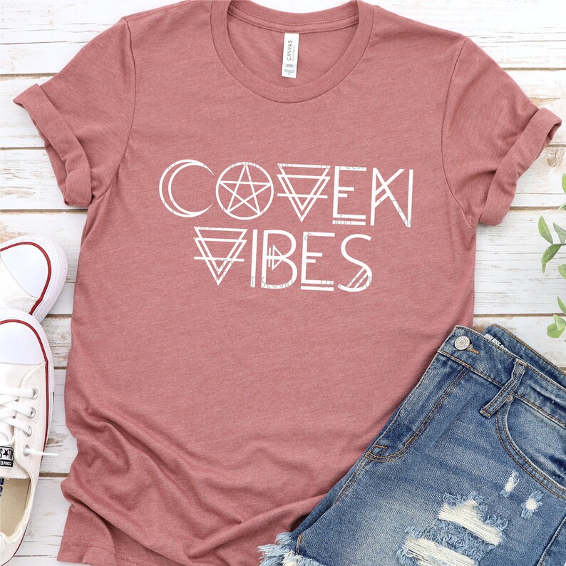 May include: A mauve t-shirt with the text "COVEN VIBES" in white. The text is stylized with geometric shapes and symbols, including a crescent moon, a pentagram, and triangles.