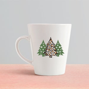 K&ouml;nnte beinhalten: Wei&szlig;e Keramik-Tasse mit einem Design aus drei Weihnachtsb&auml;umen. Der mittlere Baum hat ein Leopardenmuster, die seitlichen B&auml;ume sind gr&uuml;n mit wei&szlig;en Punkten. Die Tasse hat eine konische Form und einen gebogenen Henkel.
