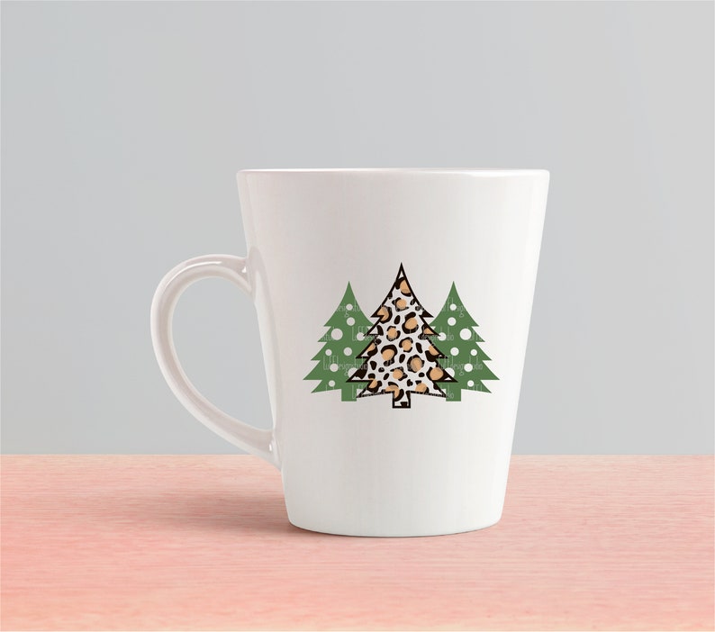K&ouml;nnte beinhalten: Wei&szlig;e Keramik-Tasse mit einem Design aus drei Weihnachtsb&auml;umen. Der mittlere Baum hat ein Leopardenmuster, die seitlichen B&auml;ume sind gr&uuml;n mit wei&szlig;en Punkten. Die Tasse hat eine konische Form und einen gebogenen Henkel.