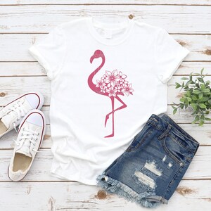May include: White t-shirt with a pink flamingo silhouette design with floral details. The flamingo is standing on one leg with its head turned to the side. The flowers are clustered around the flamingo's body.