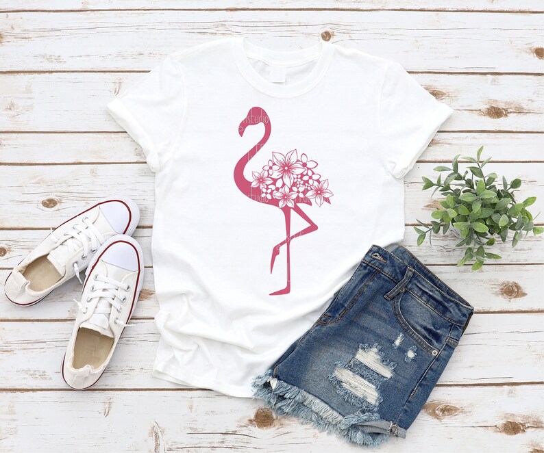 May include: White t-shirt with a pink flamingo silhouette design with floral details. The flamingo is standing on one leg with its head turned to the side. The flowers are clustered around the flamingo's body.
